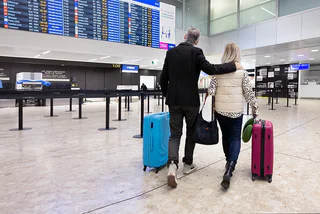 Vue d'un couple de voyageurs à l'aéroport de Genève (GVA). L'homme, portant une veste,  tient la femme par l'épaule. La femme, avec une veste clair, jean clair et sac en bandouliere marron, marche a ses cotes. Ils transportent chacun une valise cabine à roulettes vers les portes d'embarquement